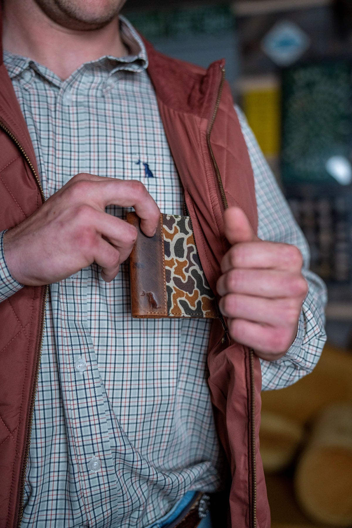 A close-up of a person wearing a rust-brown zippered vest over a green and white checkered shirt, holding a small brown leather wallet with a tan and black animal print pattern against their chest.