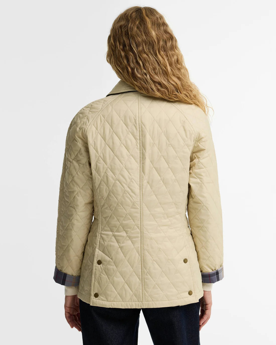 A woman viewed from the back wearing a quilted beige jacket with dark blue striped cuffs and a dark collar, paired with dark jeans, against a plain white background.
