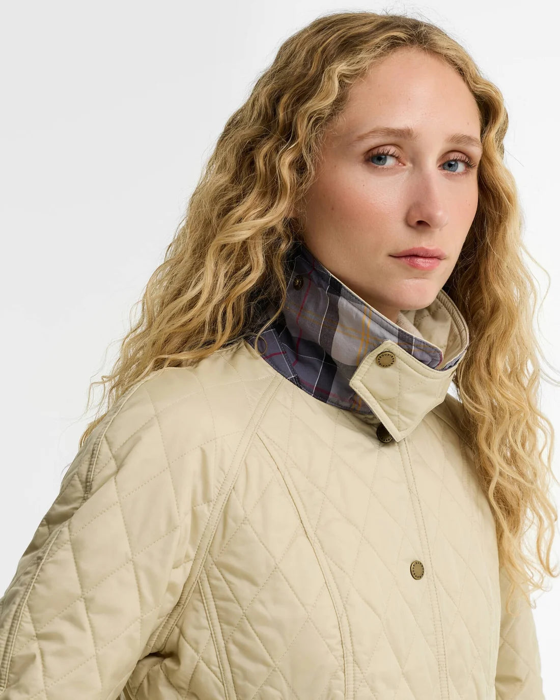 A close-up of a woman wearing a beige quilted jacket with a high plaid collar, set against a plain light gray studio background.
