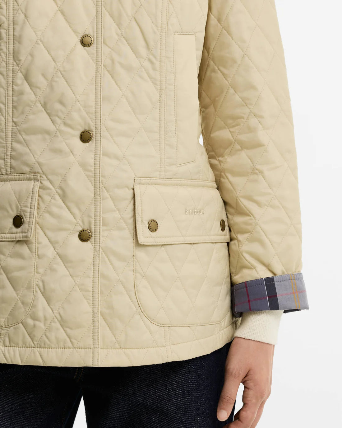 Close-up of a quilted jacket in a light beige/cream color, featuring brass-tone buttons, flap pockets, and a plaid lining visible on the turned-up cuff, worn by a model.
