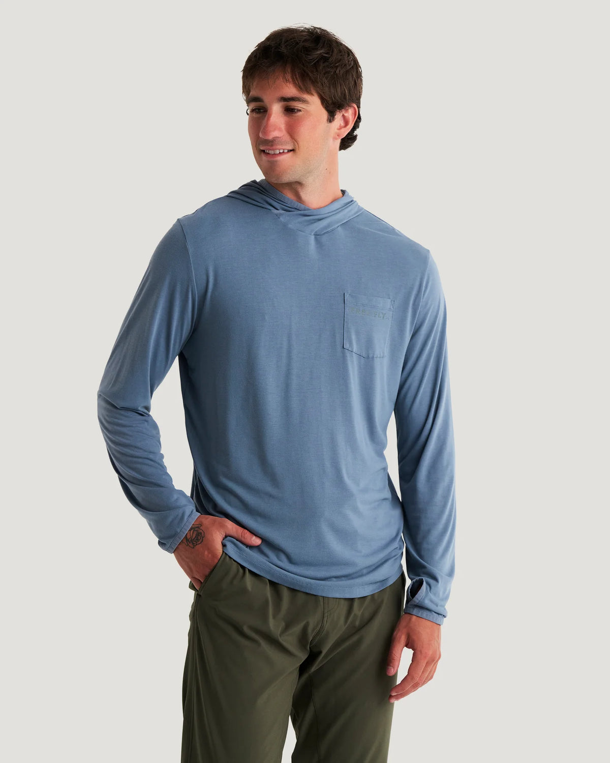 A man wearing a dusty blue long-sleeve hooded shirt with a chest pocket, paired with olive green pants, standing against a solid light gray background.
