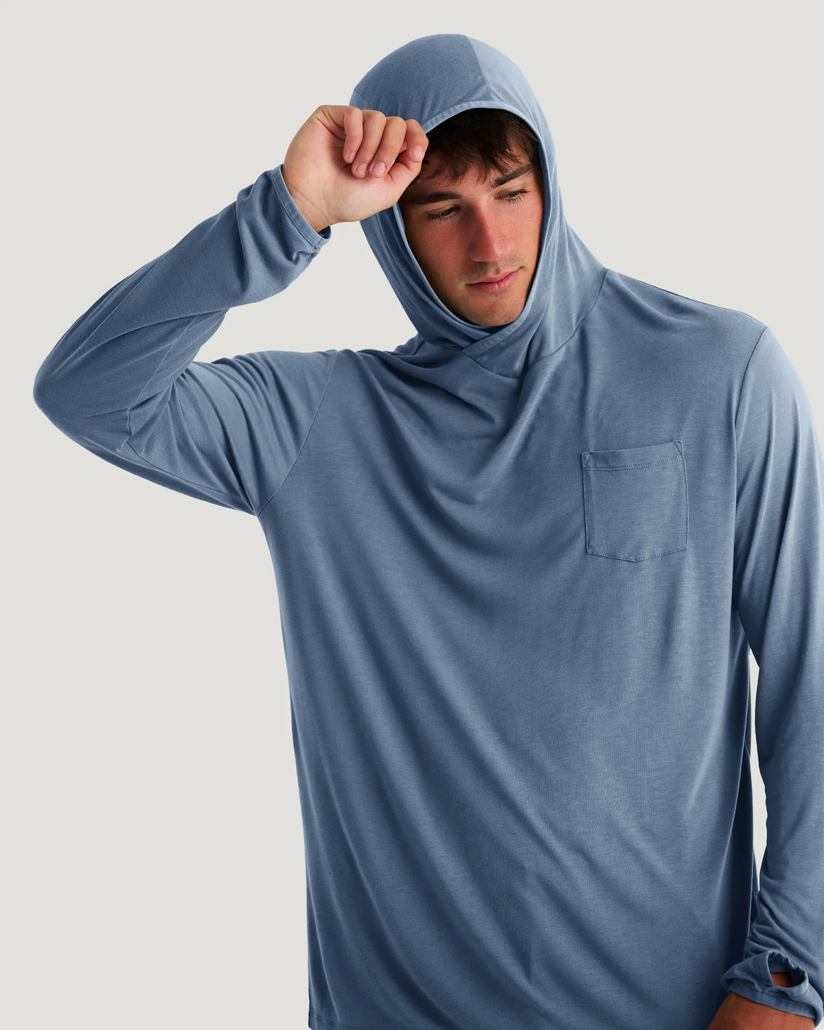 A man wearing a dusty blue long-sleeve hooded shirt with a chest pocket, posing with the hood pulled up against a light gray background.