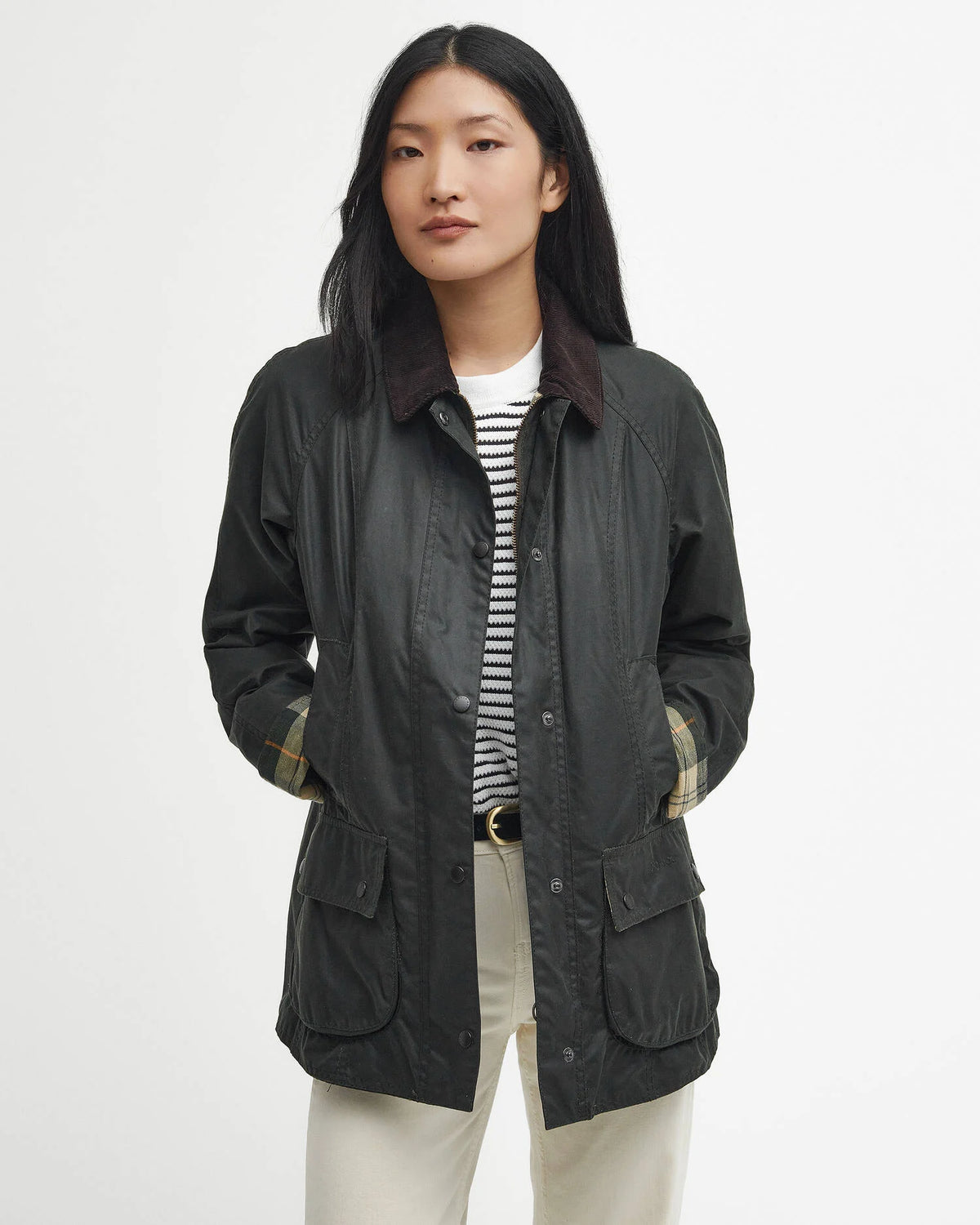 A woman modeling a dark gray waxed cotton utility jacket with a dark brown corduroy collar, worn over a black and white striped shirt and cream trousers, against a plain white studio background.