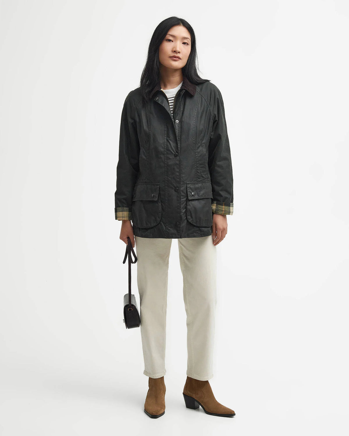 A woman stands against a plain light gray studio background wearing a dark waxed jacket over a striped shirt, off-white trousers, and brown ankle boots, while holding a small black handbag.
