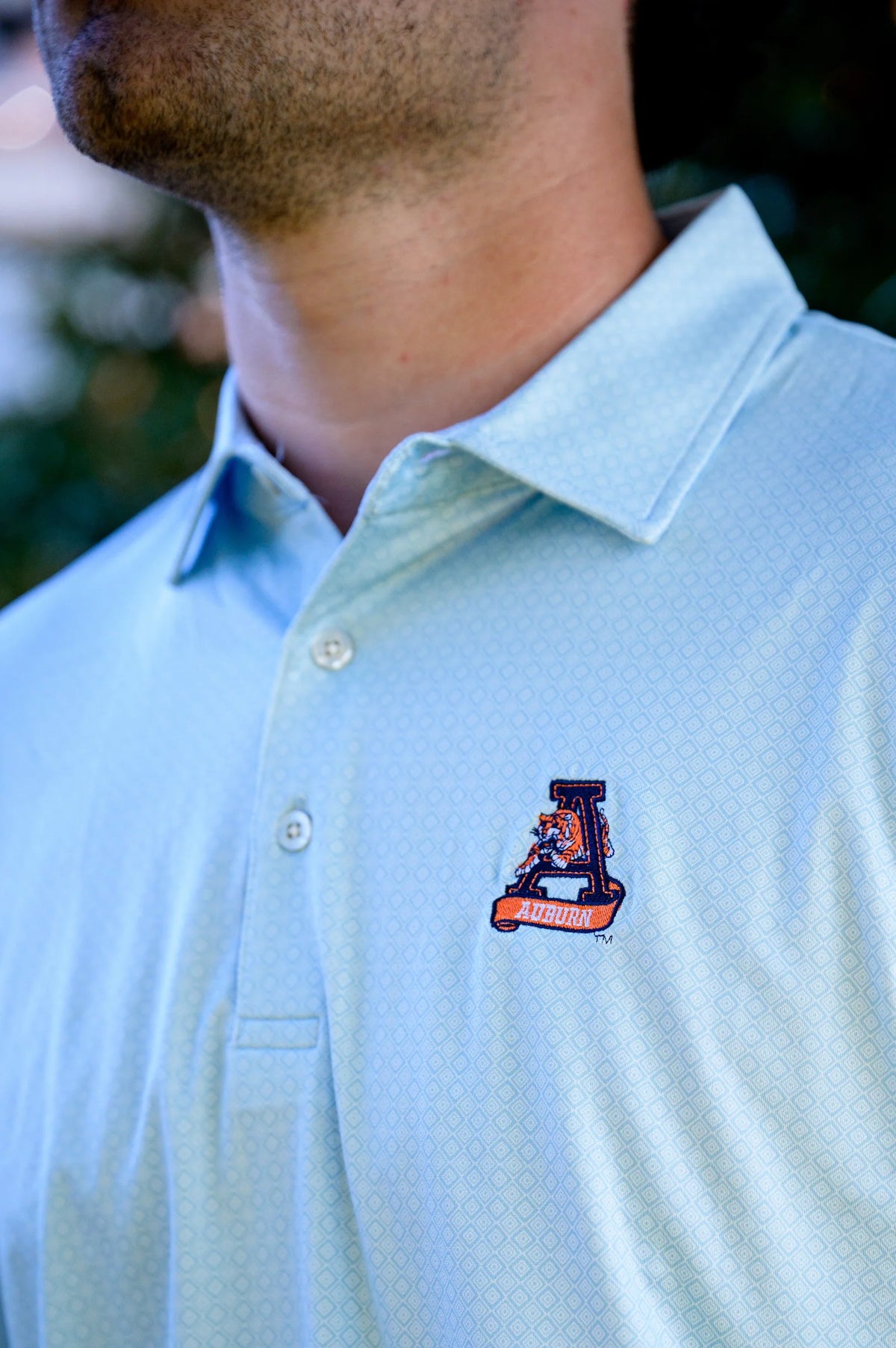 A close-up of a man wearing a light blue patterned polo shirt with a small embroidered logo on the left chest.