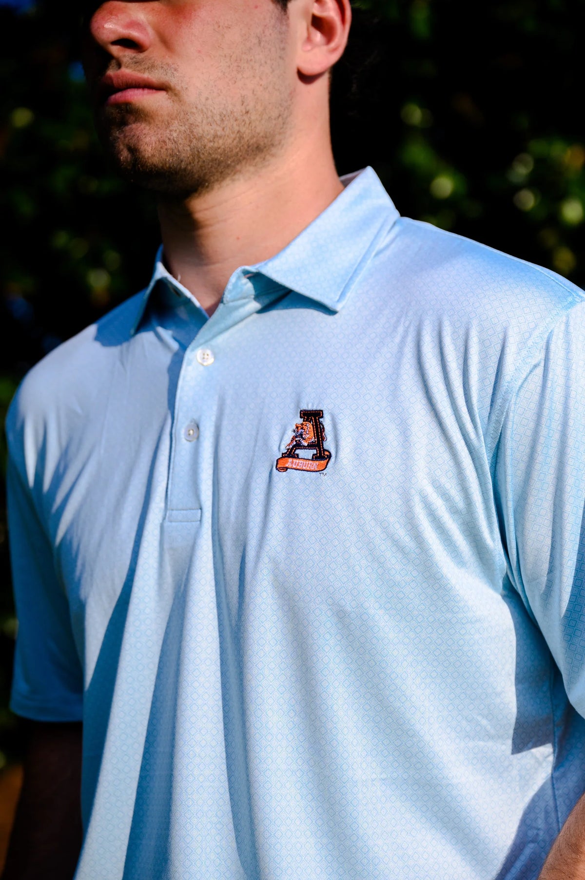 A man wearing a light blue polo shirt with a small embroidered mascot logo on the left chest, photographed outdoors with blurred green foliage in the background.