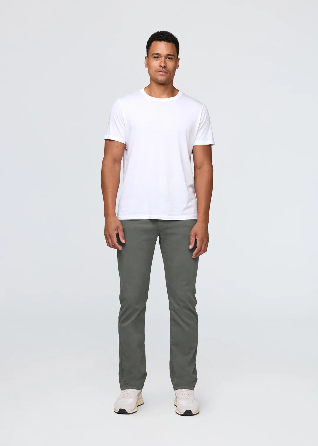 Man with dark hair wearing a white t-shirt, gray pants and white shoes standing in front of a white background. 
