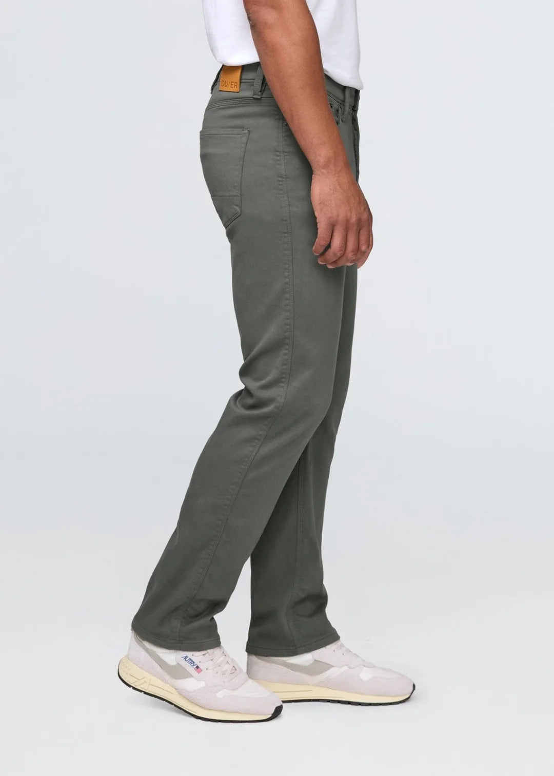 Side view of a man wearing a white t-shirt, gray pants and white shoes standing in front of a white background. 