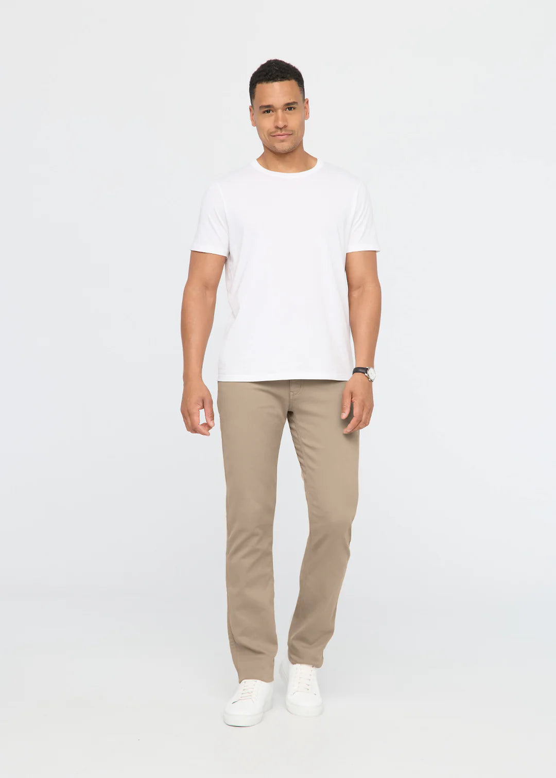 Man with dark hair wearing a white t-shirt, khaki pants and white shoes standing in front of a white background. 
