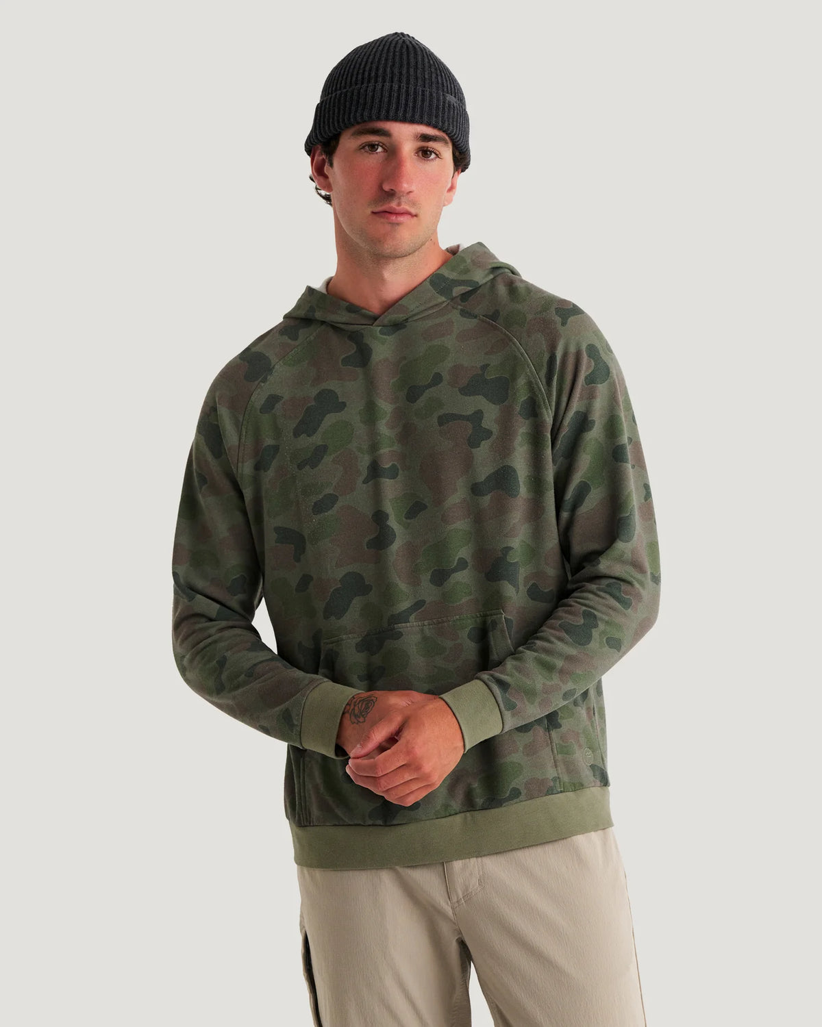 A man wearing a green camouflage pullover hoodie with raglan sleeves and a front kangaroo pocket, paired with beige pants, against a solid light gray background.