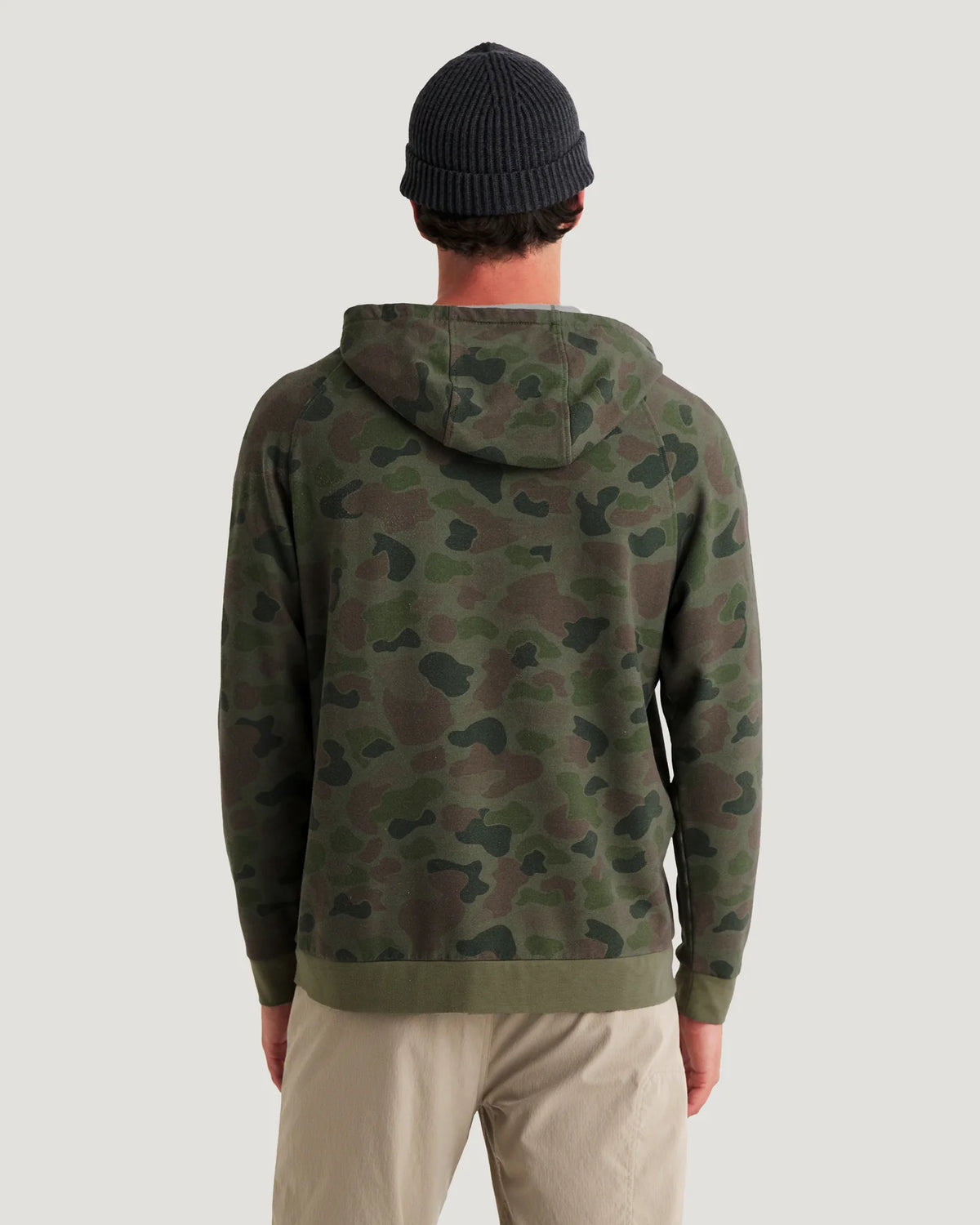 Back view of a man wearing a green camouflage pullover hoodie with raglan sleeves and a front kangaroo pocket, paired with beige pants, against a solid light gray background.