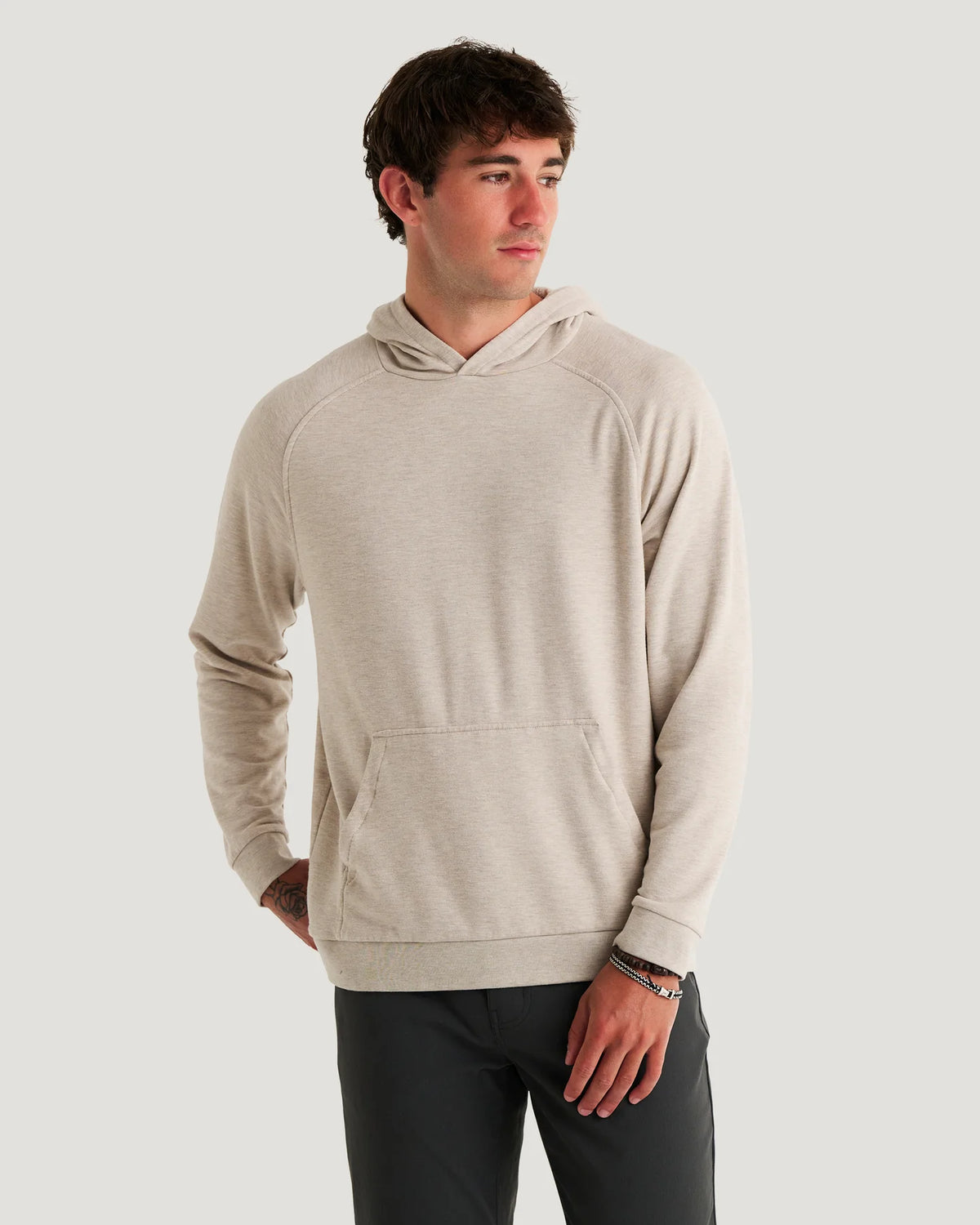 A man wearing a light beige heathered pullover hoodie with raglan sleeves and a front kangaroo pocket, paired with dark gray pants, against a solid light gray background.