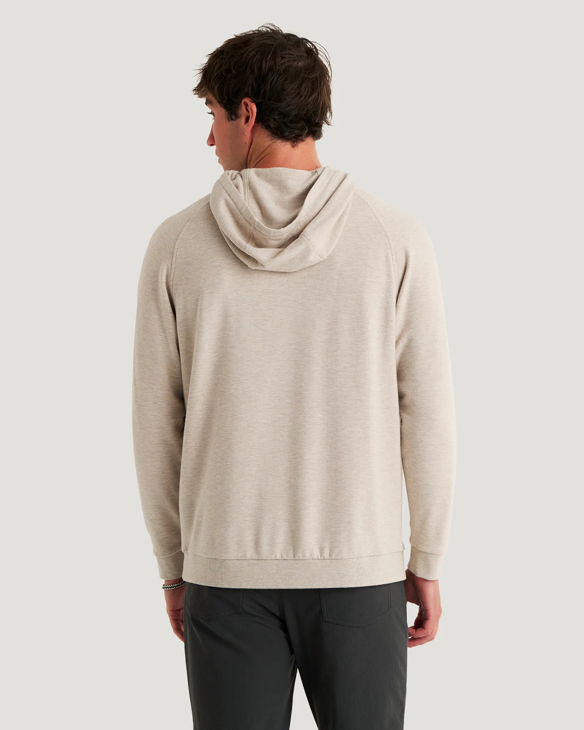 A man viewed from the back wearing a light beige hooded sweatshirt and dark gray pants, set against a solid light gray background.