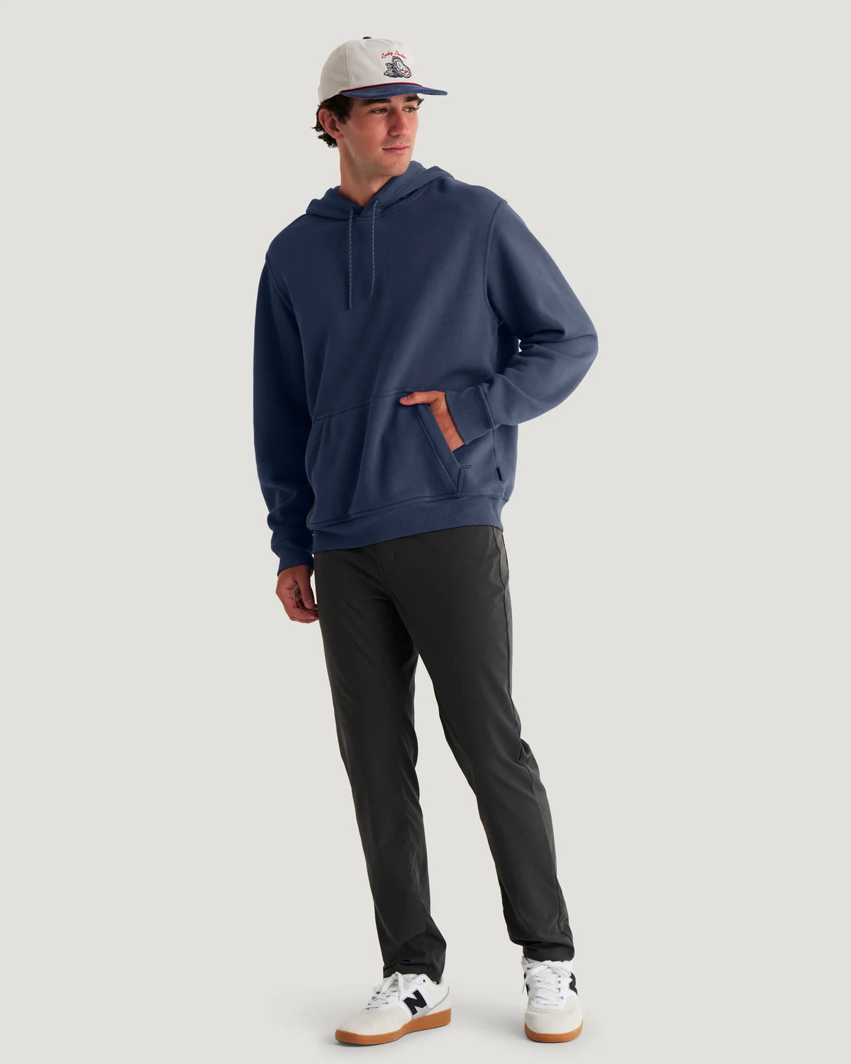 Man wearing a navy hoodie, black pants, and a cap on a light gray background