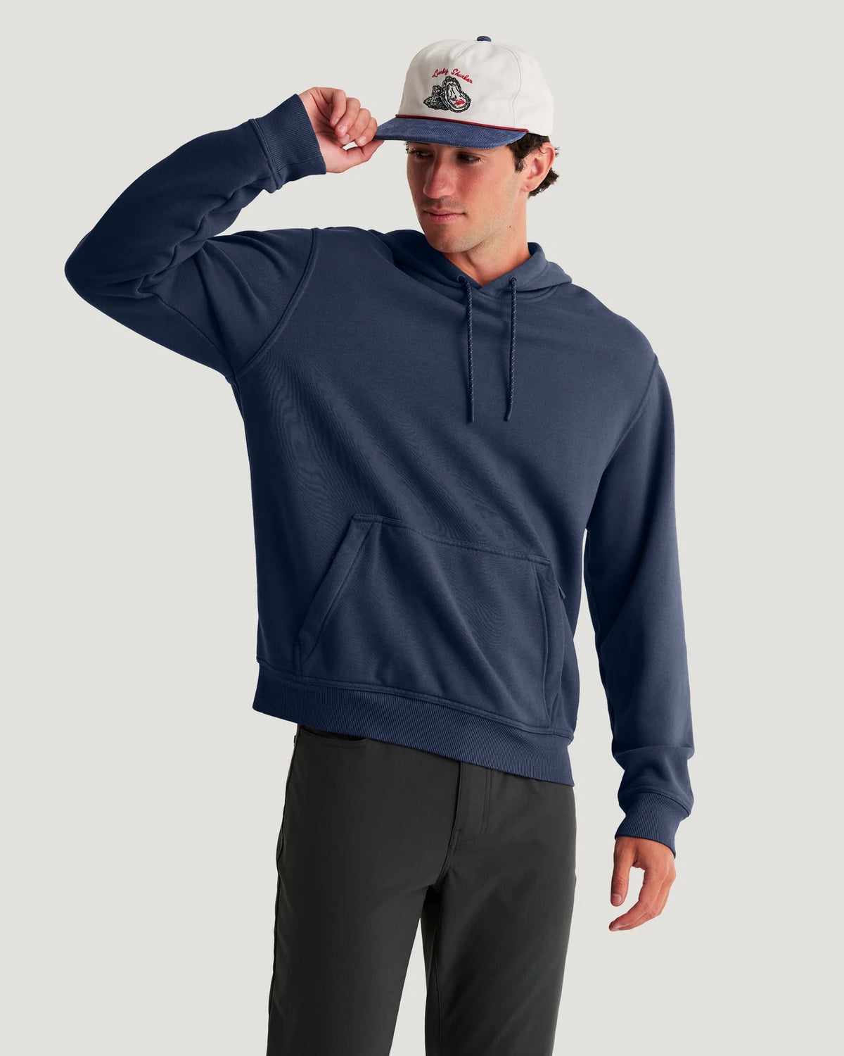 A man wearing a navy blue pullover hoodie, dark gray pants, and a white baseball cap with a navy brim, posing against a light gray studio background.