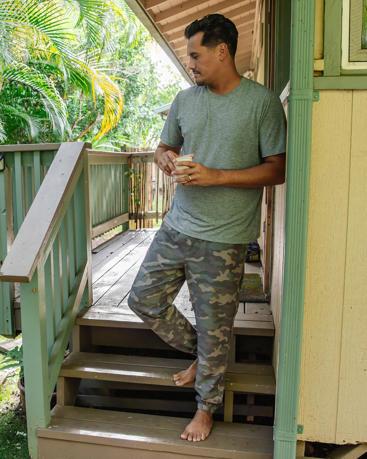 Man standing on porch steps holding a cup in his hand wearing a green t-shirt and camouflage joggers. 