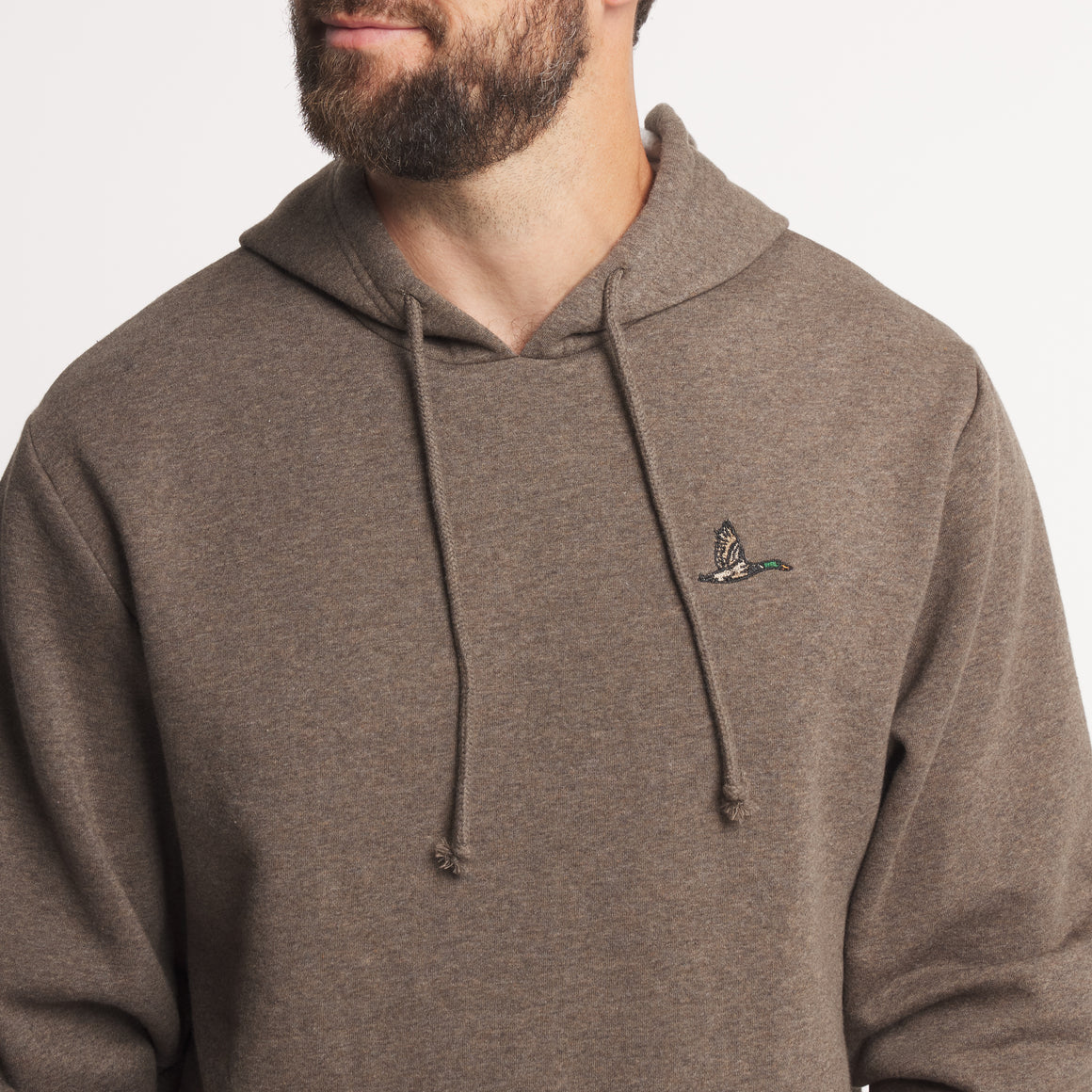 A close-up of a man wearing a brown heathered pullover hoodie with a small embroidered bird logo on the left chest, set against a plain light background.