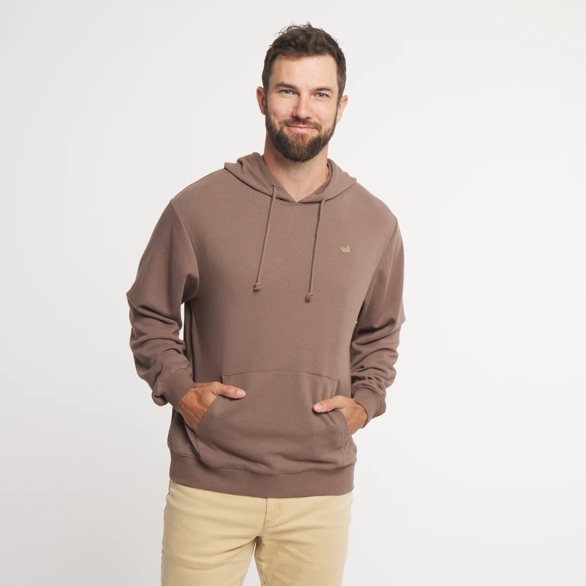 A man wearing a brown pullover hoodie with drawstrings and a kangaroo pocket, paired with beige pants, standing against a plain light gray background.
