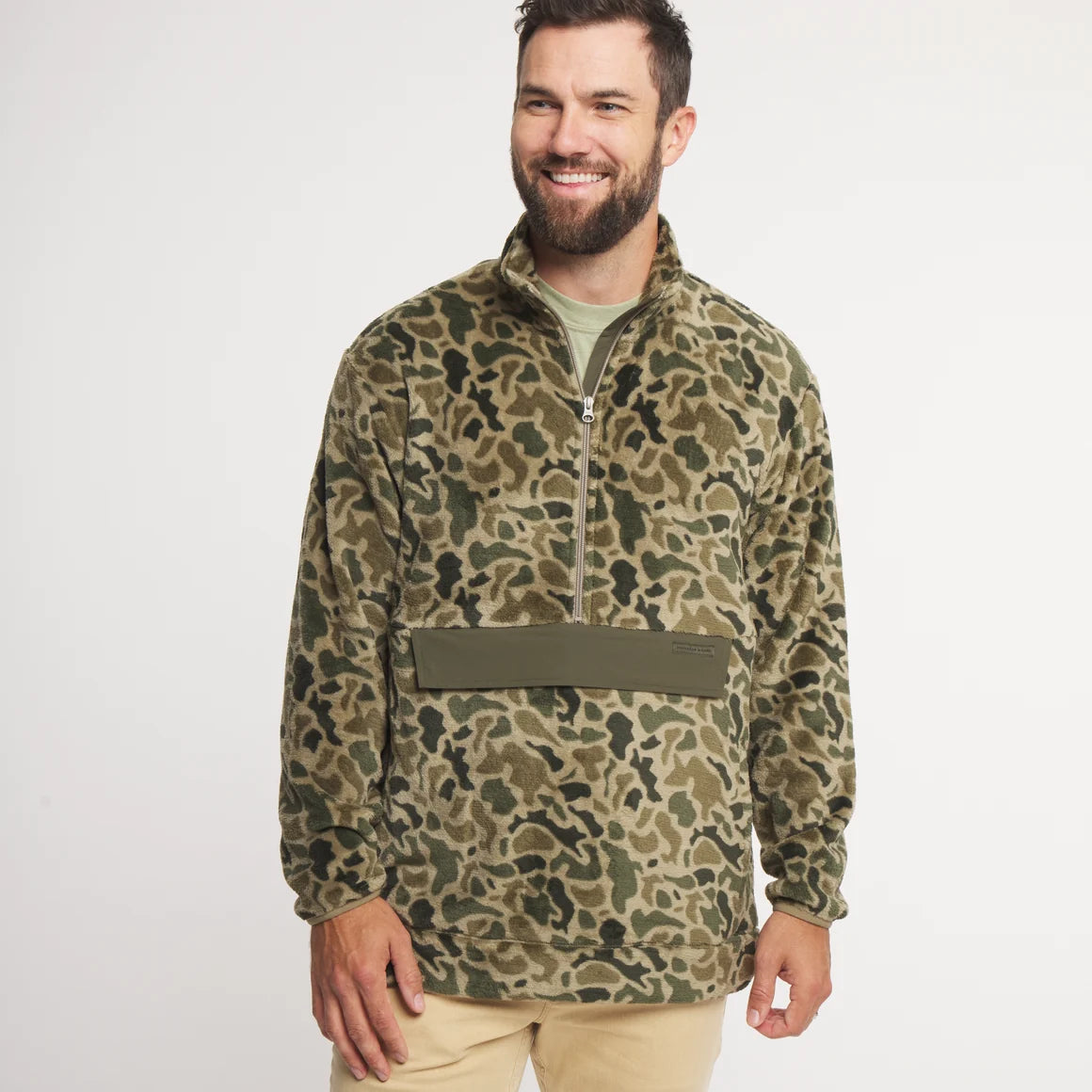 A smiling man wearing a camouflage quarter-zip fleece pullover with an olive green chest panel, paired with khaki pants, against a plain white studio background.