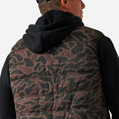 Rear view of a man wearing a quilted puffer vest with a brown and black camouflage pattern over a black hoodie, against a light gray background.