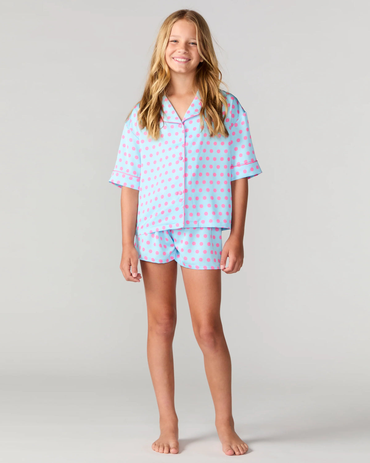 A young girl stands barefoot against a light gray studio background wearing a light blue pajama set with pink polka dots and pink piping details.