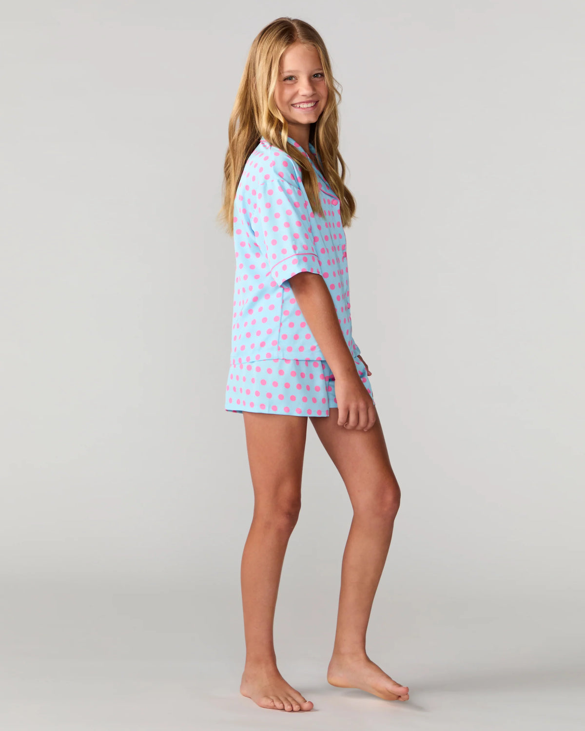 A young girl with long blonde hair stands barefoot against a light gray studio background, wearing a short-sleeve pajama shirt and matching shorts in light blue with a pink pattern.