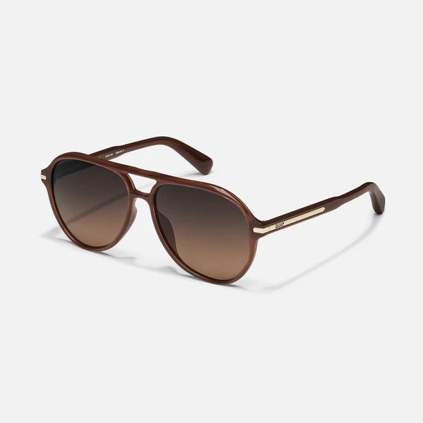 A pair of aviator-style sunglasses with a glossy brown acetate frame and gradient brown lenses, featuring a thin gold metal accent bar on the bridge and temples, set against a light gray background.