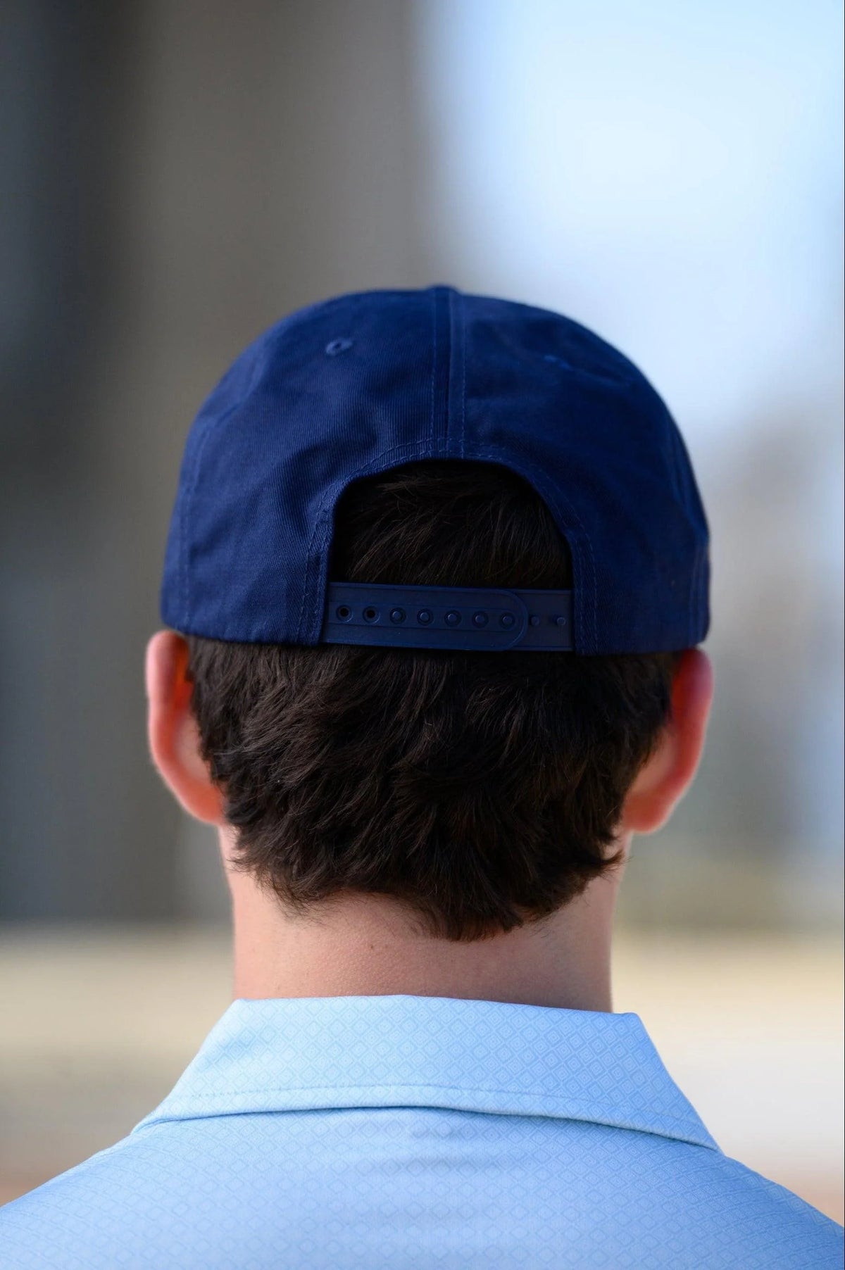 A man viewed from behind wearing a navy blue baseball cap and a light blue collared shirt, set against a softly blurred outdoor background.