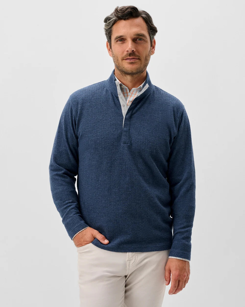 A man wearing a textured navy blue long-sleeve quarter-placket sweater over a collared shirt and light beige trousers, standing against a light gray studio background.