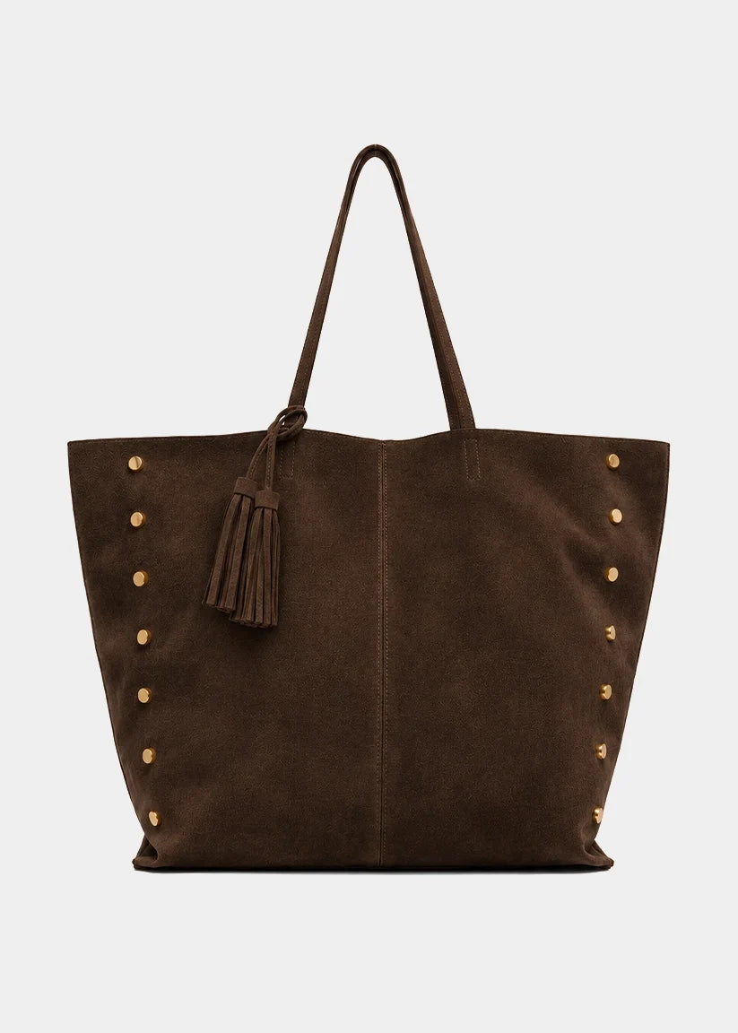 A dark brown suede tote bag with gold-tone metal studs and a matching brown leather tassel charm, shown against a light gray background.