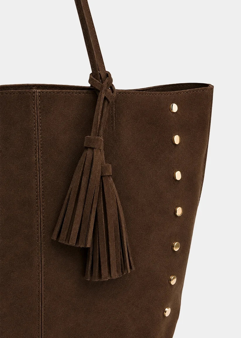 A close-up of a dark brown suede tote bag featuring a matching tassel charm and gold-tone metal studs along the side, set against a light gray background.