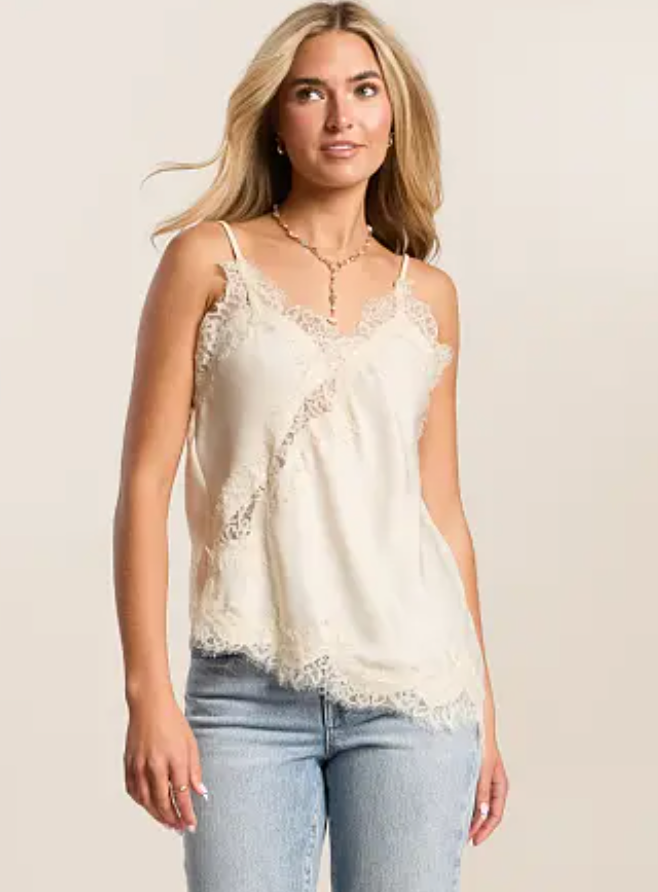 A woman modeling a cream-colored lace-trimmed camisole top paired with light-wash blue jeans against a solid light beige background.
