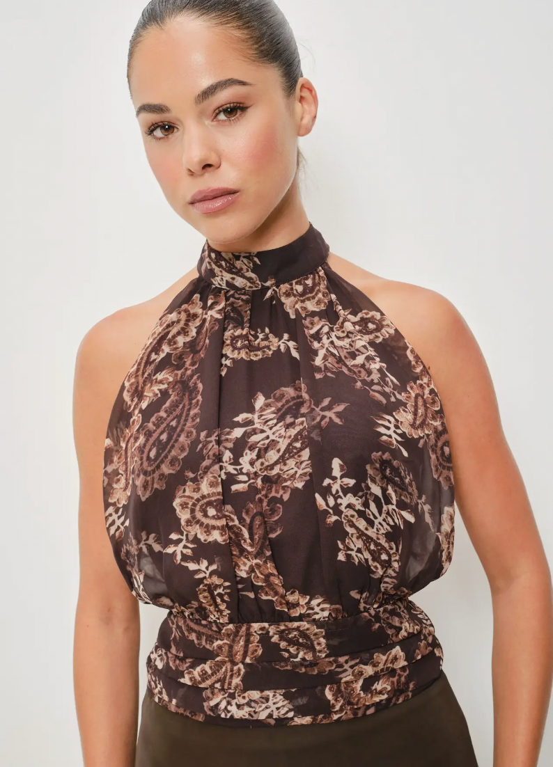 A woman wearing a sheer halter-neck top with a brown and beige paisley pattern, paired with dark brown bottoms, photographed against a plain light gray studio background.