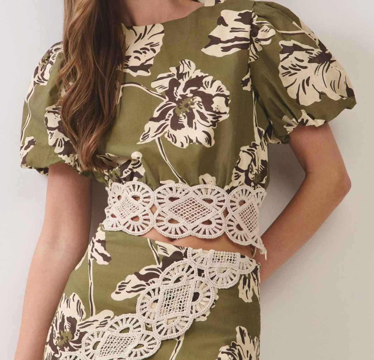 A woman wearing a two-piece olive green floral set with cream and brown hibiscus flowers and a wide cream lace waistband and skirt hem.