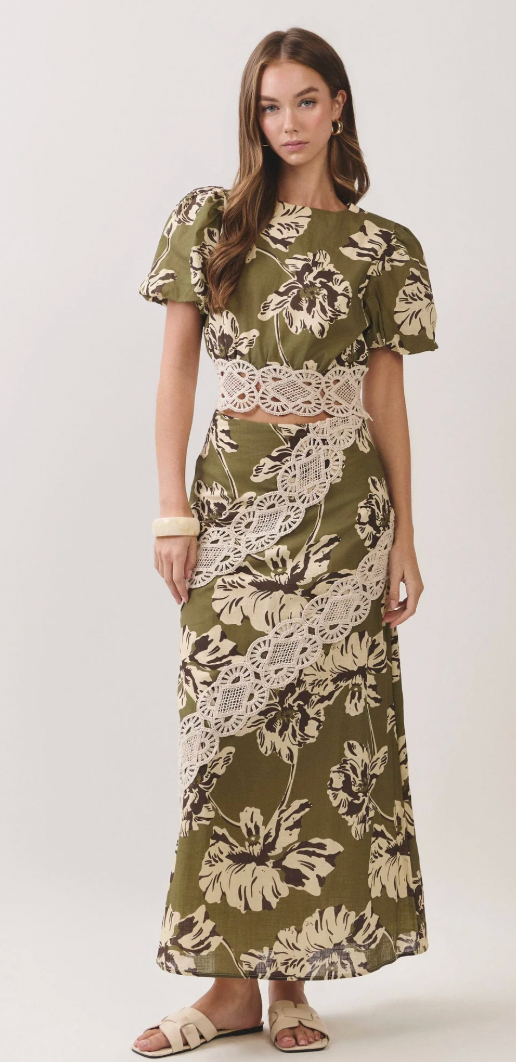 A woman modeling a two-piece olive green floral set with beige and brown tones, featuring a short-sleeve crop top and a matching maxi skirt with lace trim, against a plain light background.