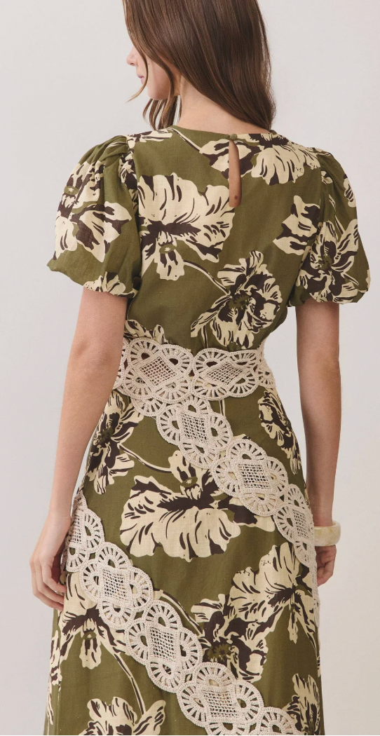 A woman shown from the back wearing an olive green dress with a large cream and dark brown floral print and a white lace applique band at the waist, against a plain light background.