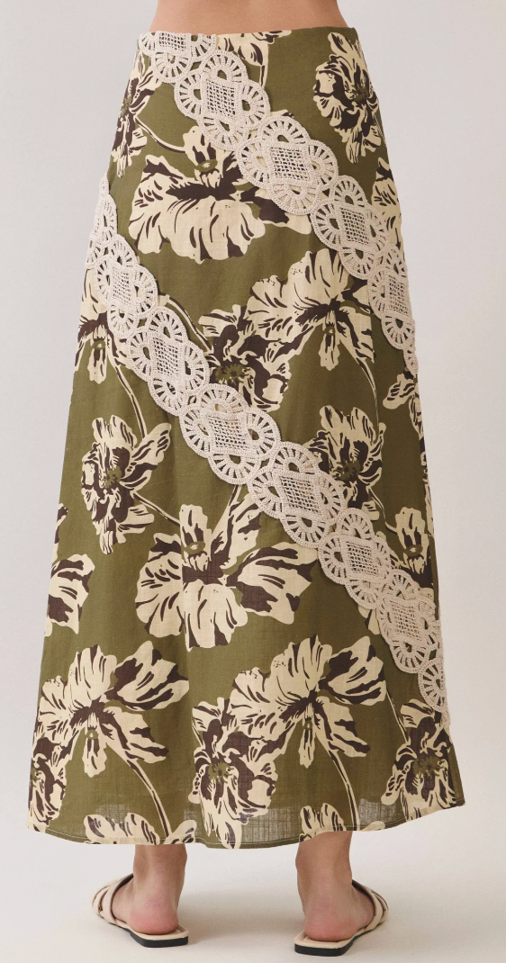 A woman wearing a long olive green skirt with a cream and dark brown floral print and a diagonal lace overlay, paired with beige slide sandals, photographed against a light gray studio background.