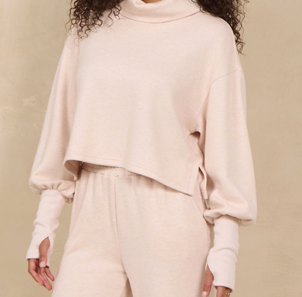 A person wearing a light blush pink knit loungewear set, featuring a cropped turtleneck sweater with long balloon sleeves and matching sweatpants, against a neutral beige background.