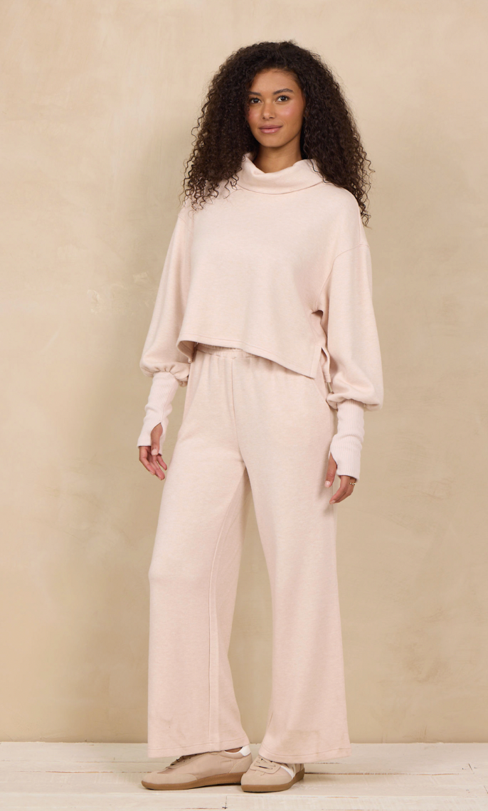 A woman modeling a light blush pink two-piece lounge set consisting of a cropped long-sleeve turtleneck top and matching wide-leg pants, paired with beige flat shoes against a textured beige studio backdrop.
