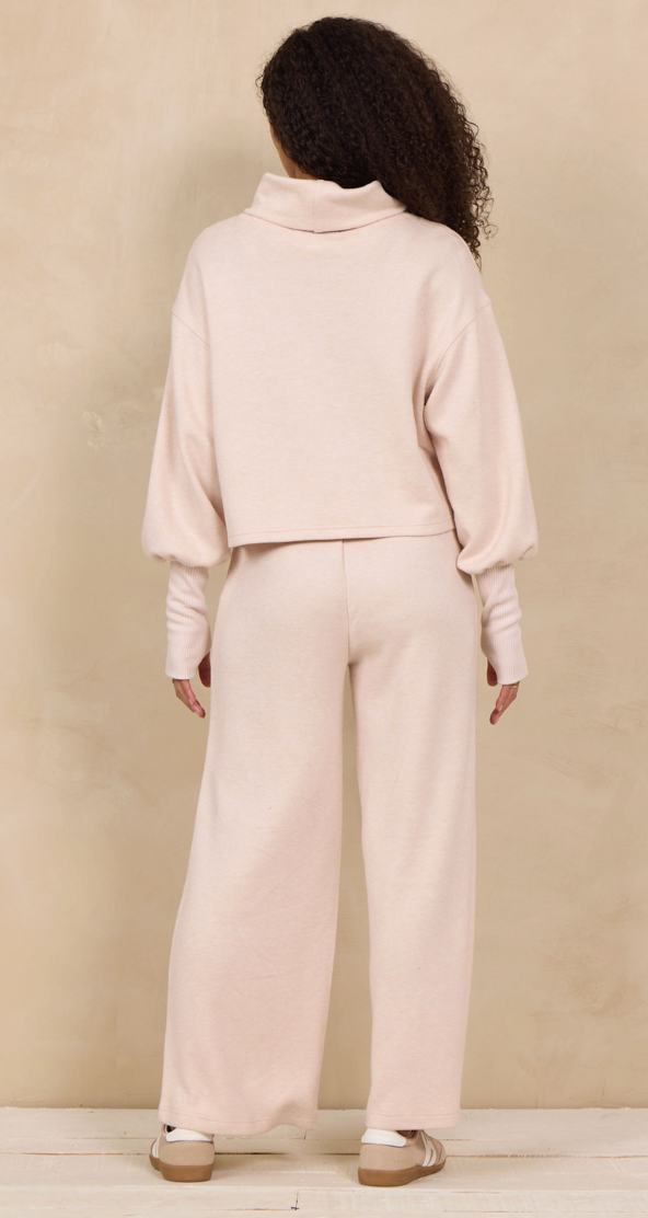 A model viewed from the back wearing a light blush pink two-piece lounge set consisting of a cropped long-sleeve turtleneck top and matching wide-leg pants, paired with beige flat shoes against a textured beige wall.