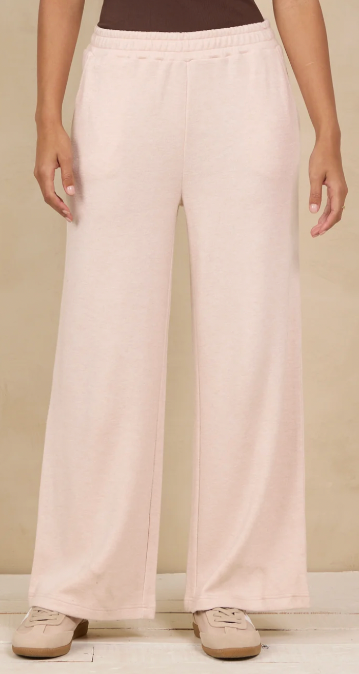 A person wearing light blush pink wide-leg sweatpants with an elastic waistband and side pockets, paired with beige sneakers, against a warm beige studio background.