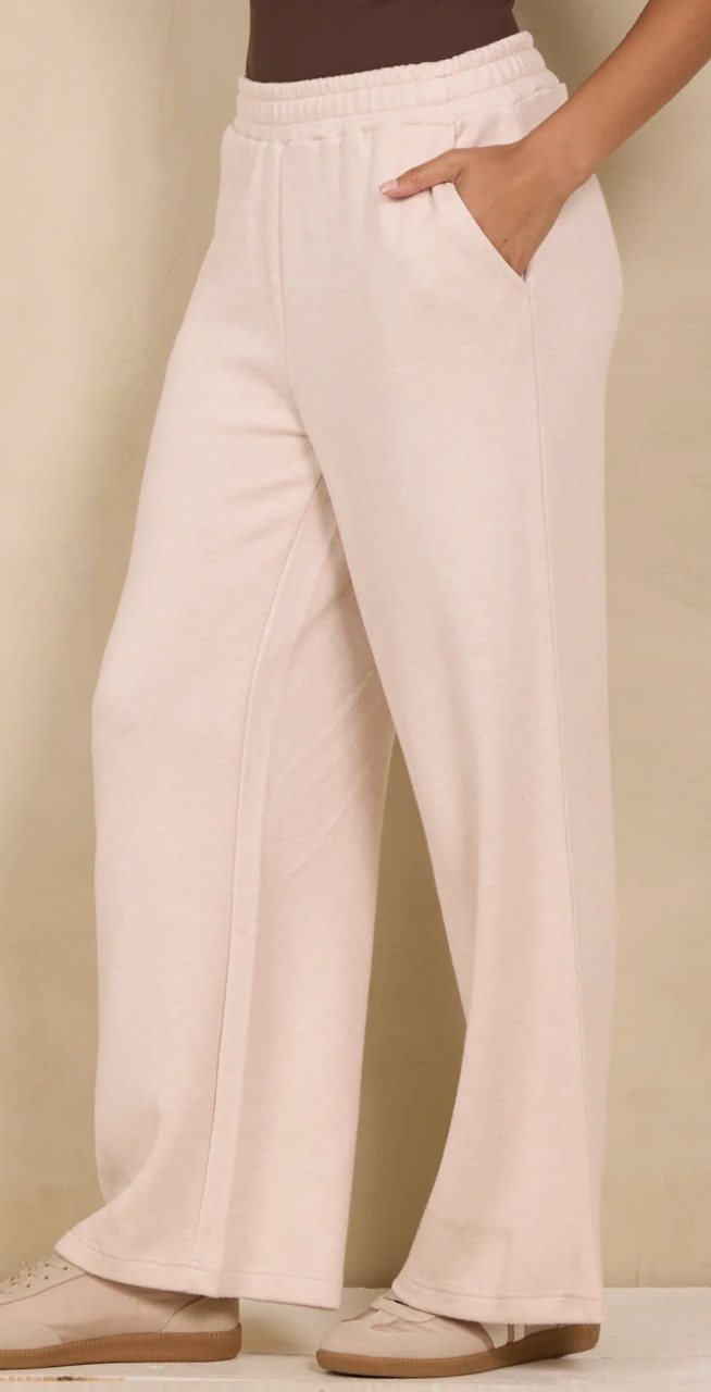 A person wearing light blush pink wide-leg sweatpants with an elastic waistband and side pockets, paired with beige sneakers, against a warm beige studio background.