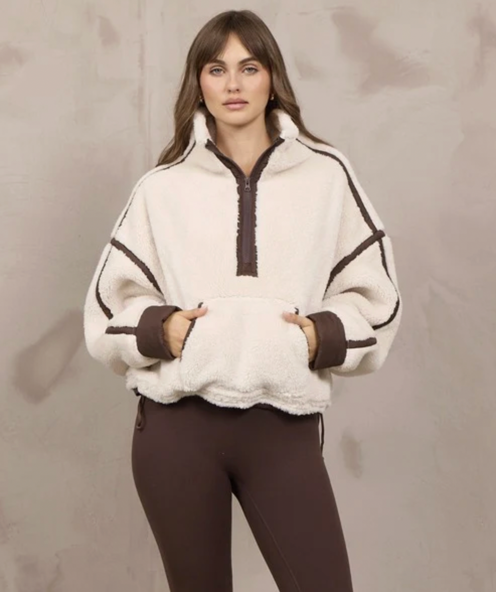 A woman modeling a cream sherpa half-zip sweatshirt with dark brown trim and matching brown leggings, standing against a textured light gray wall.