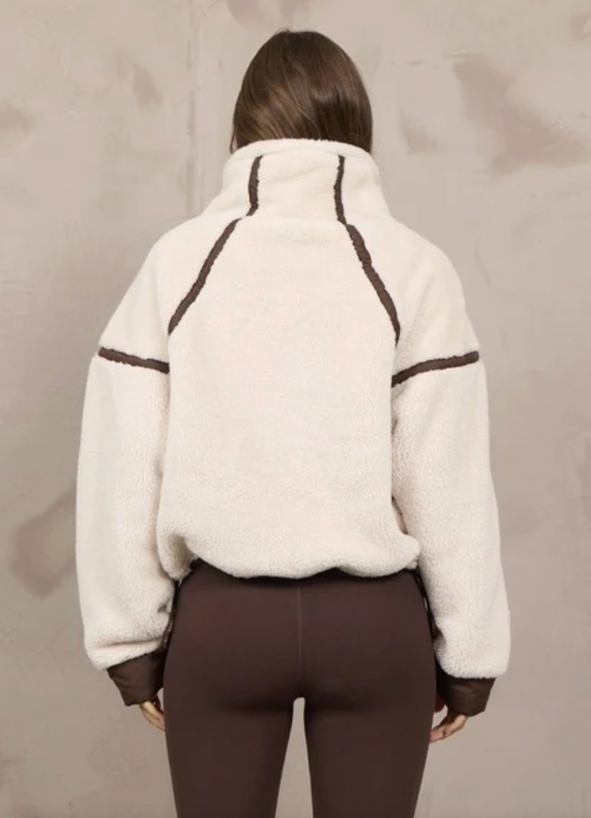 A person shown from the back wearing a cream sherpa fleece pullover with dark brown contrast stitching and matching brown cuffs, paired with brown leggings, against a textured light gray wall.
