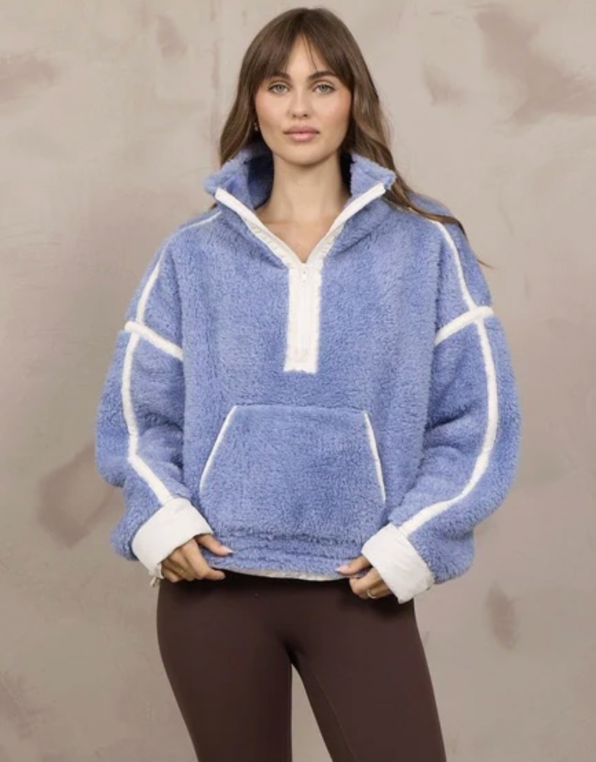 A woman modeling a light blue sherpa fleece half-zip pullover sweatshirt with white contrast stitching and trim, paired with dark brown leggings, against a textured beige wall.