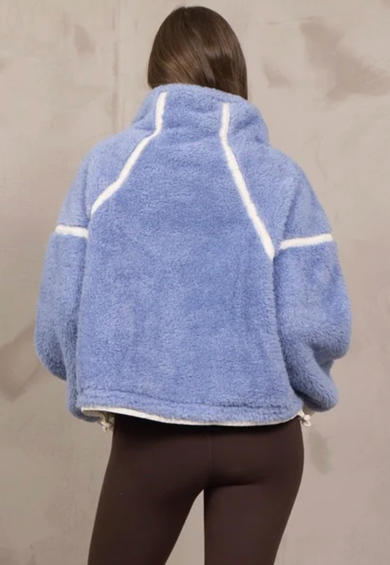 A person shown from the back wearing a light blue fuzzy cropped jacket with white contrast stitching details, paired with dark brown leggings, against a neutral beige wall.