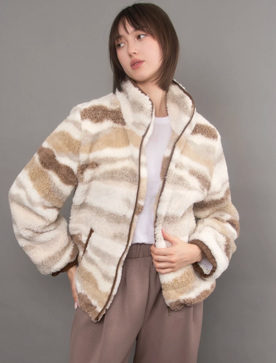 Model wearing a plush ivory, tan, and taupe striped zip-up jacket with a high collar, paired with a white top and brown pants.
