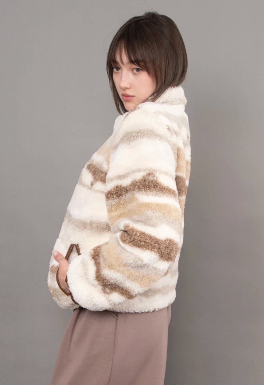 Model wearing a plush ivory, tan, and taupe striped zip-up jacket with a high collar, paired with a white top and brown pants.