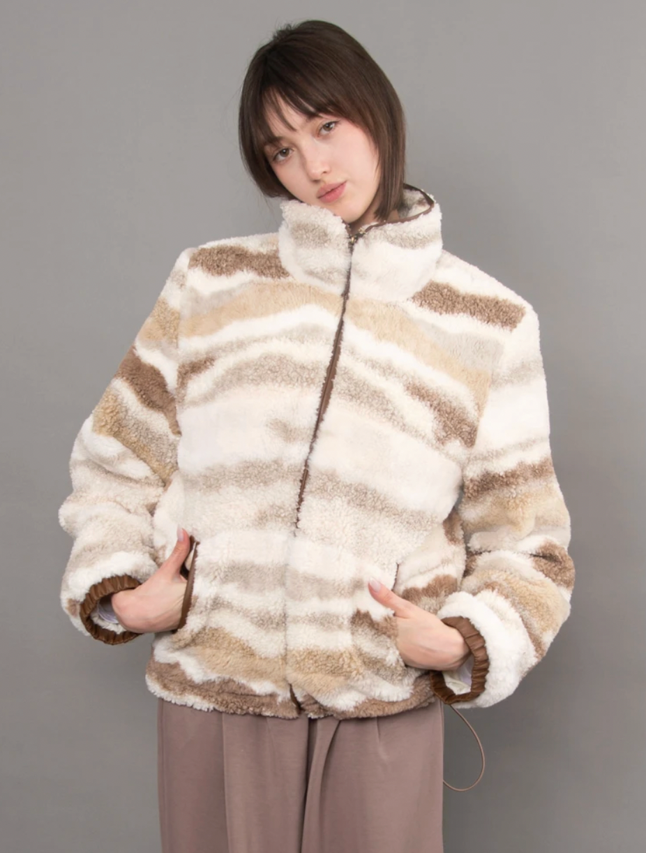 Model wearing a plush ivory, tan, and taupe striped zip-up jacket with a high collar, paired with a white top and brown pants.