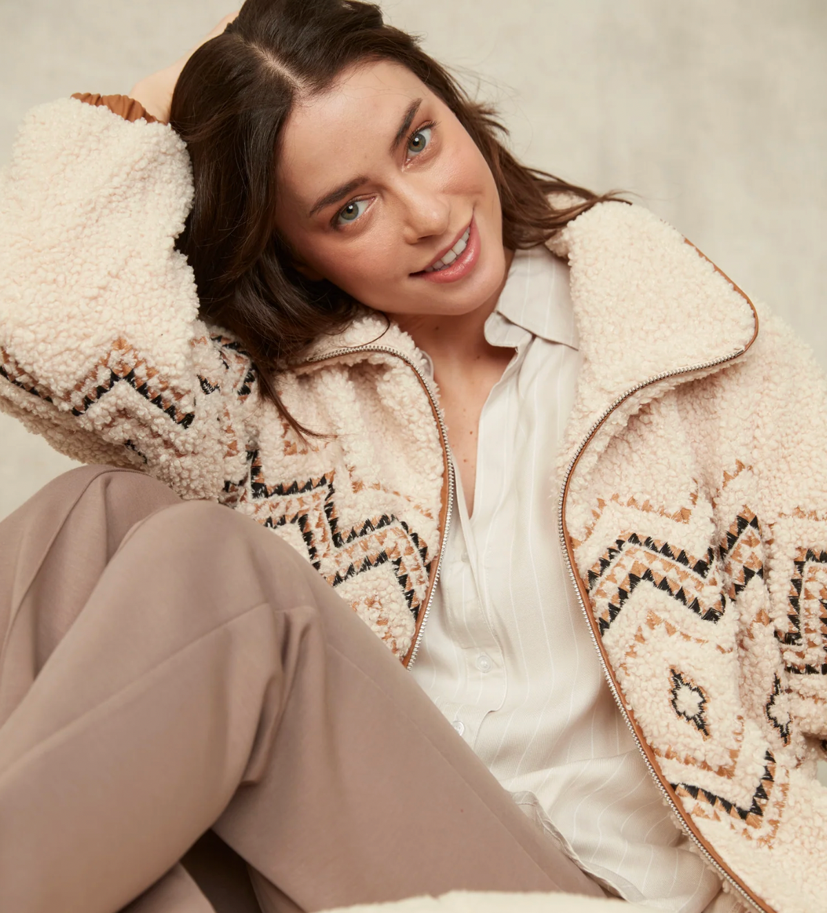 A woman wearing a cream sherpa zip-up jacket with a brown and black geometric pattern, styled over a white shirt and beige trousers, set against a neutral textured background.